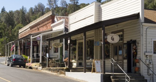 A quaint street lined with historic buildings and shops, surrounded by trees and a clear blue sky.