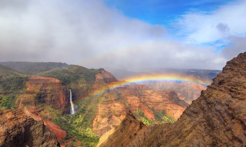 A vibrant rainbow arcs over lush green valleys and a waterfall amidst rugged, colorful cliffs under a cloudy sky.