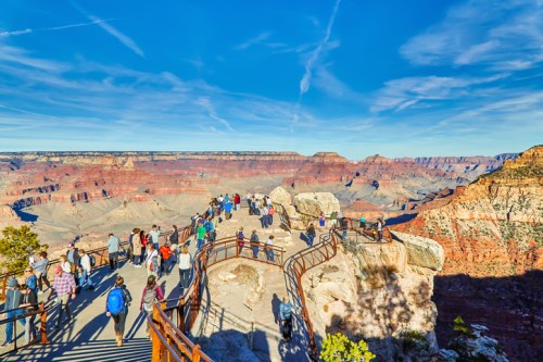 A crowded viewpoint at the Grand Canyon, showcasing vast rock formations and a clear blue sky.
