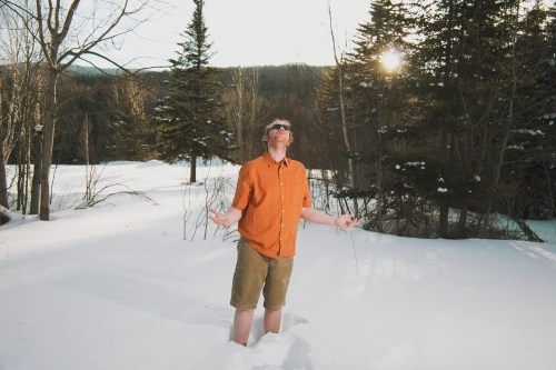 A man in a short-sleeve shirt and shorts stands in deep snow, looking up with arms outstretched, surrounded by trees.