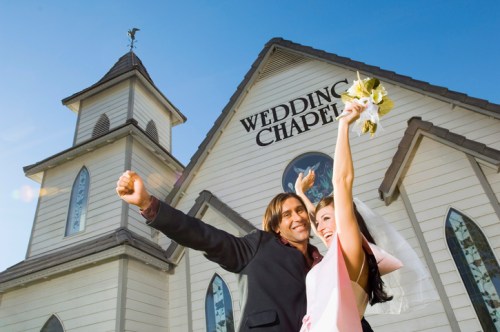A happy couple celebrates outside a wedding chapel, with the bride raising her bouquet and the groom cheering.