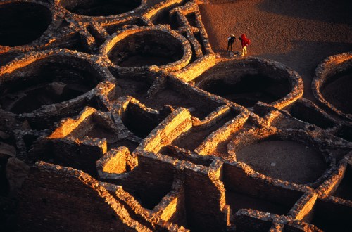 Aerial view of ancient ruins with circular structures, two people exploring the site in warm, golden light.