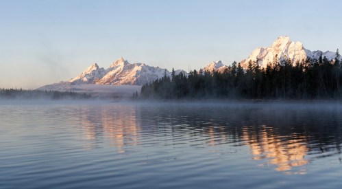 Serene lake reflecting snow-capped mountains at sunrise, surrounded by mist and evergreen trees.