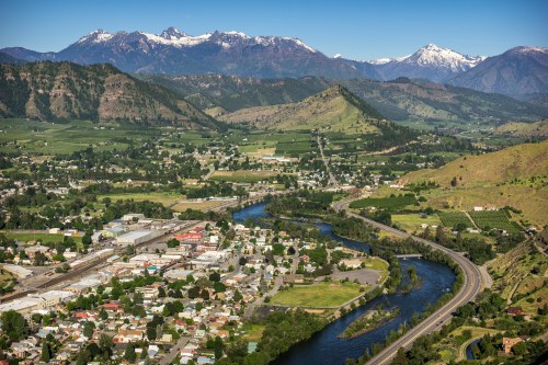 Aerial view of a valley with a river, town, and mountains in the background, featuring lush greenery and snow-capped peaks.