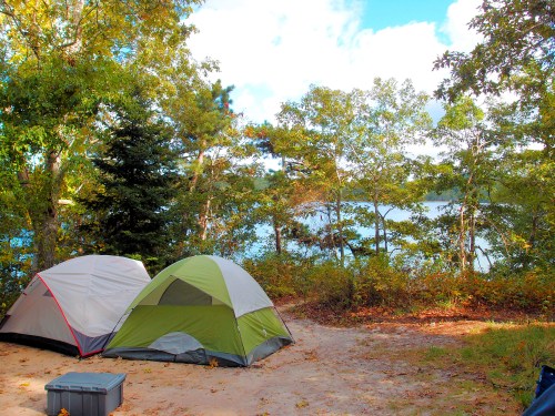 Two tents set up by a serene lakeside, surrounded by trees and a clear blue sky.