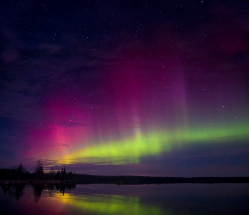 A vibrant display of the northern lights with pink and green hues reflecting on a calm lake under a starry sky.