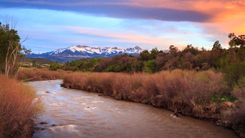 A serene river flows through a landscape of trees and shrubs, with snow-capped mountains under a colorful sky.