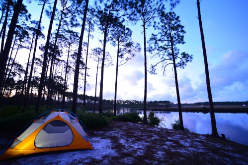 A tent by a calm lake surrounded by tall trees at dusk, with soft light illuminating the tent.