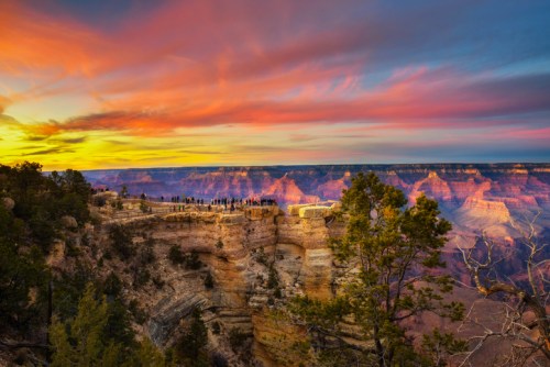 A vibrant sunset over the Grand Canyon, with silhouettes of people on the rim and colorful clouds in the sky.