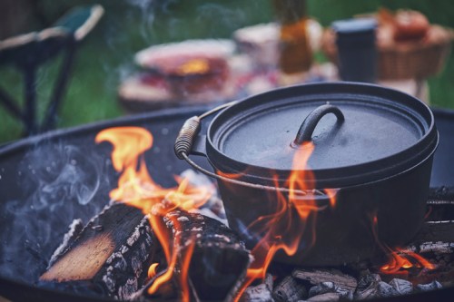 A cast iron pot sits over a campfire, surrounded by flames and logs, with food and drinks in the background.