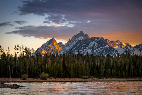 Sunset over rugged mountains reflecting in a calm lake, surrounded by lush forests and a colorful sky.