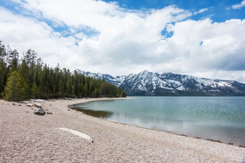 A serene lakeside scene with a pebbly shore, surrounded by lush trees and snow-capped mountains under a cloudy sky.