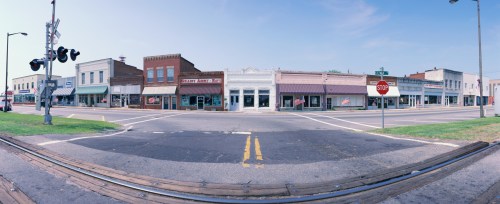A panoramic view of a small town street with shops, American flags, and a railroad crossing.