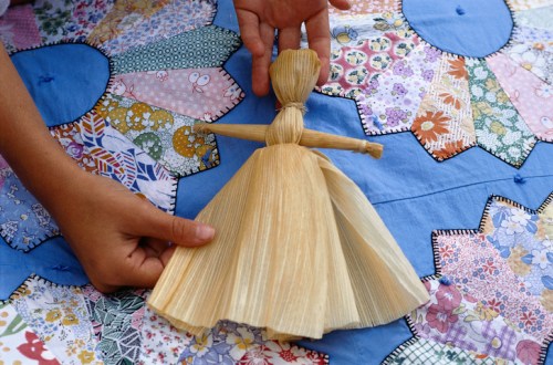 A person holds a handmade corn husk doll over a colorful quilt with various patterns.
