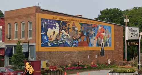 Colorful mural on a brick building depicting various scenes and figures, with a sign for "Village Floral" nearby.