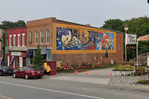 A colorful mural on a brick building, depicting various figures and scenes, alongside a street with parked cars.