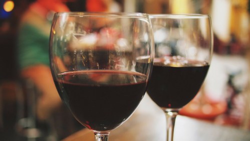Two glasses of red wine on a table, with a blurred background of people and warm lighting.