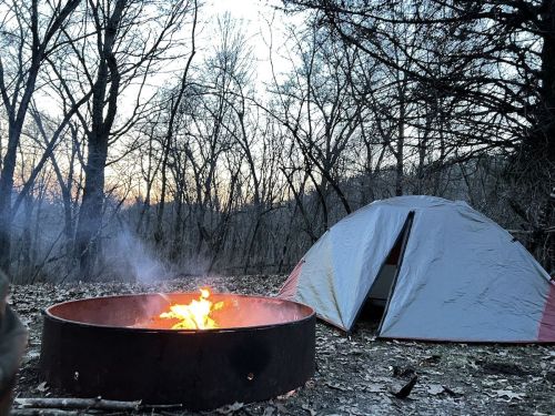 A campsite with a glowing campfire and a tent, surrounded by trees at dusk.