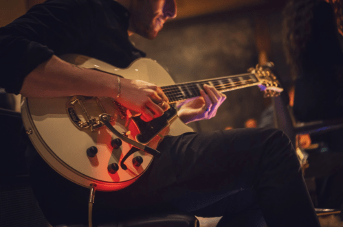 A musician plays a white electric guitar, seated in a dimly lit setting, focused on their performance.