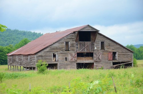 A weathered wooden barn with a rusty metal roof, set against a backdrop of green hills and cloudy skies.