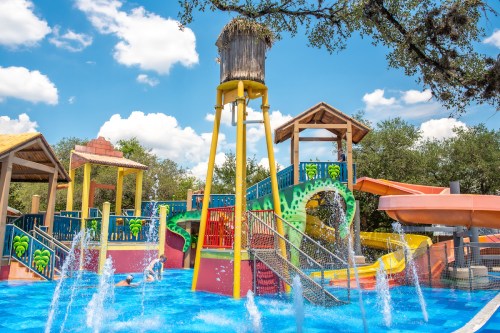 Colorful water park with slides, splash features, and a large water tower under a blue sky with fluffy clouds.