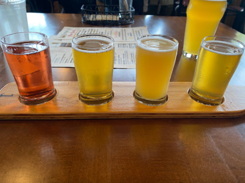 A wooden flight paddle holds four small glasses of different colored beers on a table.