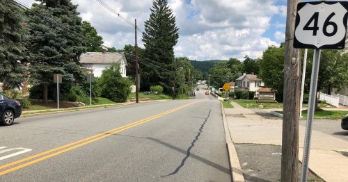 A view of a quiet road with a highway sign for Route 46, lined with trees and houses under a partly cloudy sky.