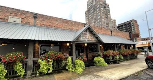 A brick restaurant with a metal roof, adorned with hanging plants, set against a city skyline.