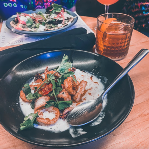 A black bowl with sliced grilled octopus, cilantro, and a creamy sauce, alongside a cocktail and a plate of salad.