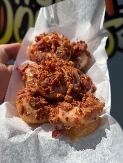 A hand holding a tray of mini donuts topped with bacon bits and a creamy glaze, set against a colorful background.