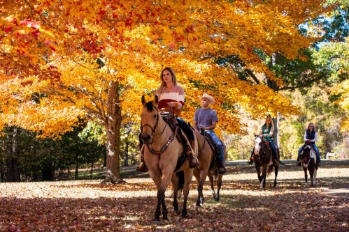 A woman and a child ride horses through a vibrant autumn landscape with colorful leaves.