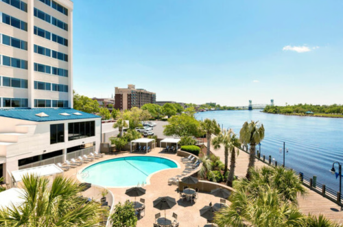 A sunny view of a hotel pool area beside a river, with palm trees and buildings in the background.