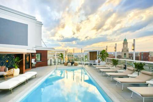 Rooftop pool with lounge chairs, surrounded by city views and a dramatic sunset sky.