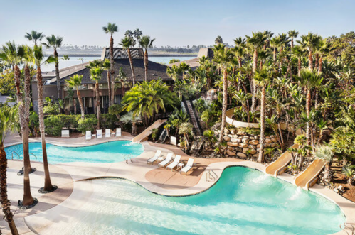 A scenic pool area with slides, surrounded by palm trees and lush greenery, overlooking a calm body of water.