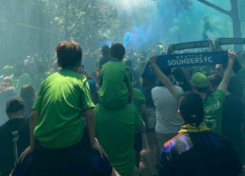 A crowd of fans in green shirts, some holding scarves, celebrating with smoke in the air during a Seattle Sounders FC event.