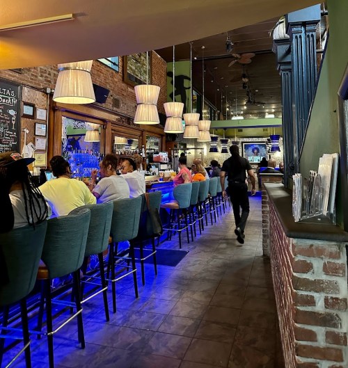 A lively bar scene with patrons seated at a long counter, illuminated by pendant lights and a vibrant blue glow.