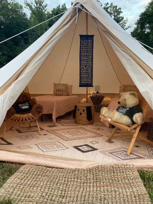 Cozy glamping tent with plush bears, a bed, and decorative signs promoting positivity, surrounded by nature.