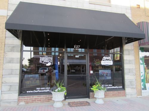 Front view of a storefront with a black awning, large windows, and planters on a brick sidewalk.