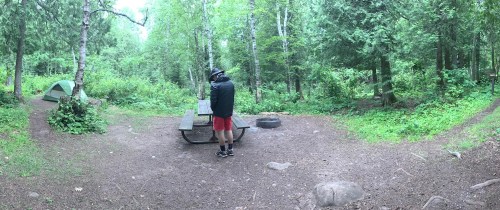 A person in a black jacket and red shorts stands by a picnic table in a wooded campsite with a tent nearby.