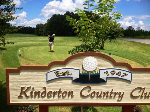 A golfer swings on a lush green course near the Kinderton Country Club sign, established in 1947.