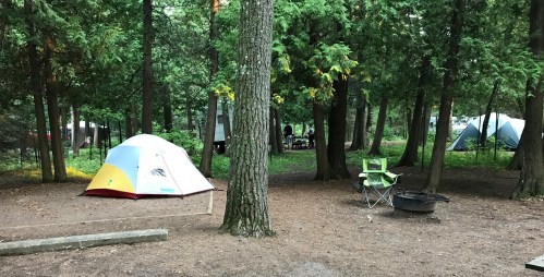 A campsite in a forest with a colorful tent, a campfire ring, and a folding chair among tall trees.