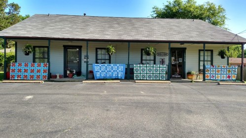 A small building with a gray roof, decorated with colorful quilts and potted plants on either side.
