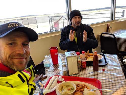 Two men sitting at a table in a diner, enjoying meals with a view of an airfield outside.