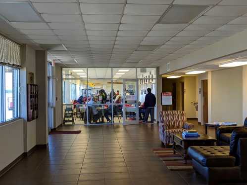 A spacious hallway leading to a glass-walled room with people gathered around tables, discussing or working together.