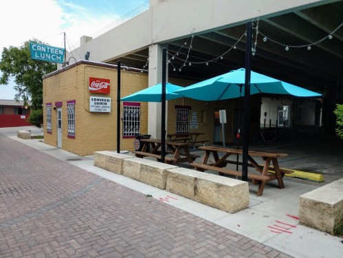 A small eatery with outdoor seating under blue umbrellas, located beneath a structure with string lights.