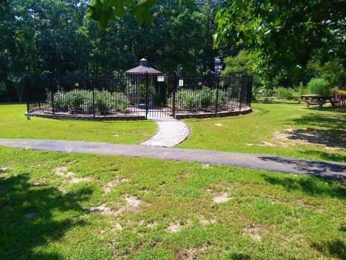 A circular garden enclosed by a black fence, featuring a gazebo and a stone path, surrounded by green grass and trees.