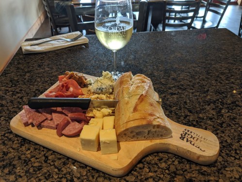 A wooden board with assorted cheeses, cured meats, bread, and a glass of white wine on a dark table.