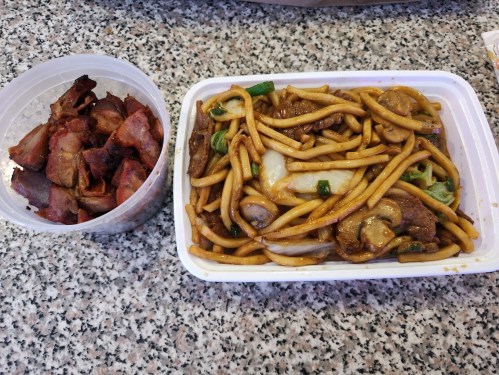 A container of sliced roasted meat next to a dish of lo mein with vegetables and beef on a speckled countertop.