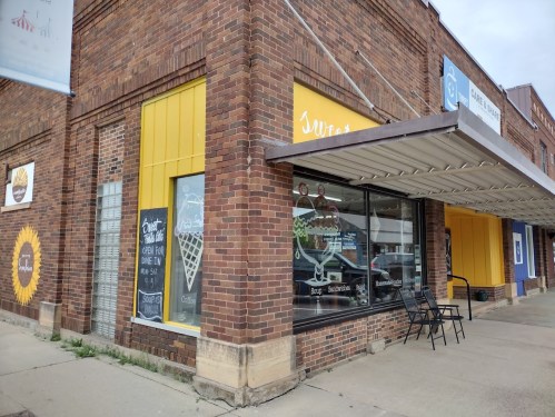 A brick building with yellow accents and large windows displaying a dessert shop's signage and outdoor seating.