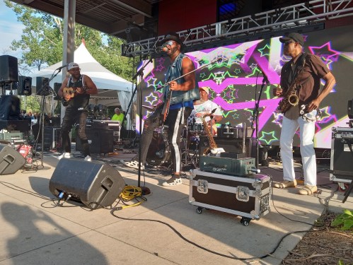 A lively band performs on stage with colorful visuals, featuring musicians on guitar, drums, and saxophone.